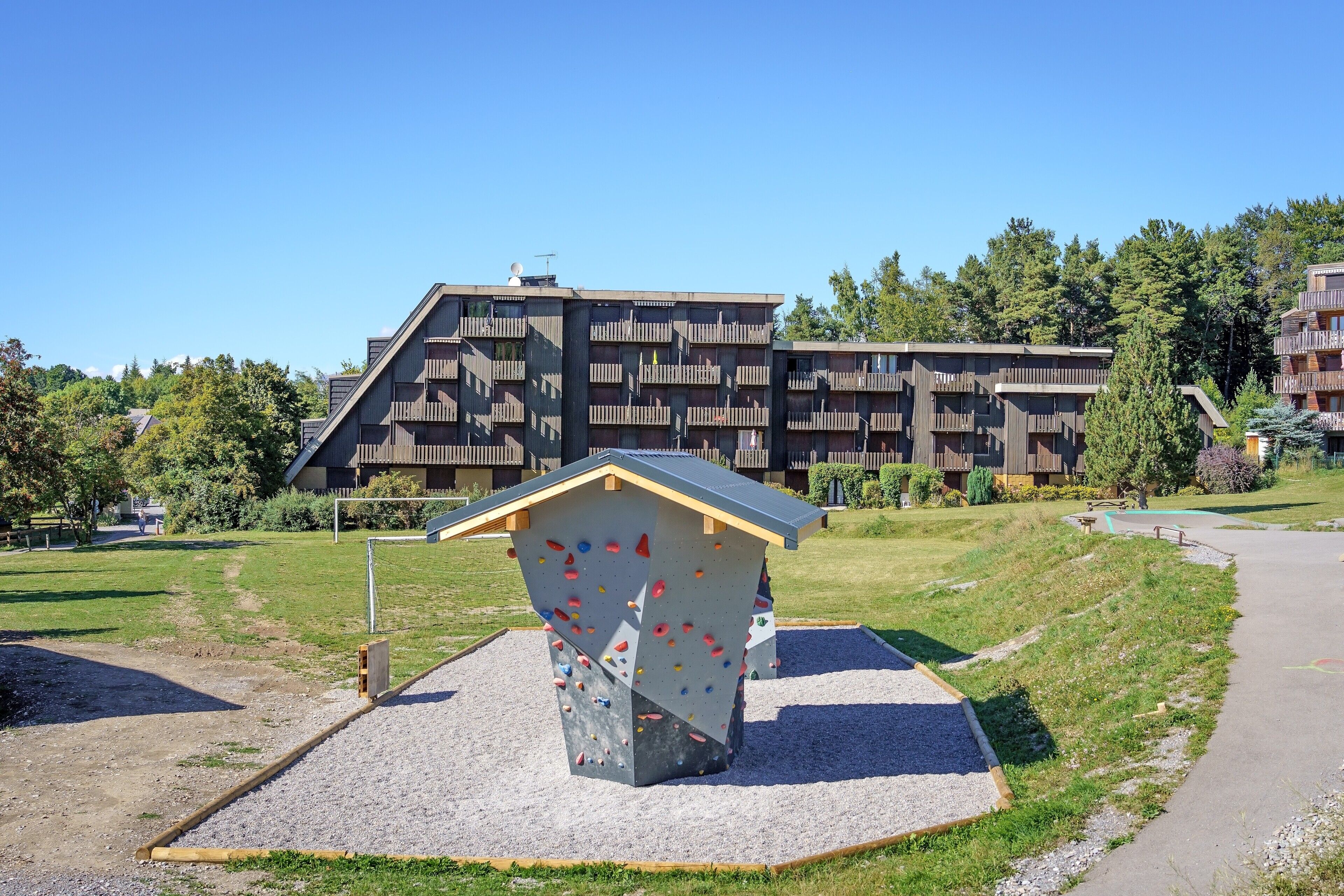 A climbing wall with a wooden exterior and a gravel base, set against a backdrop of modern buildings and a clear sky.