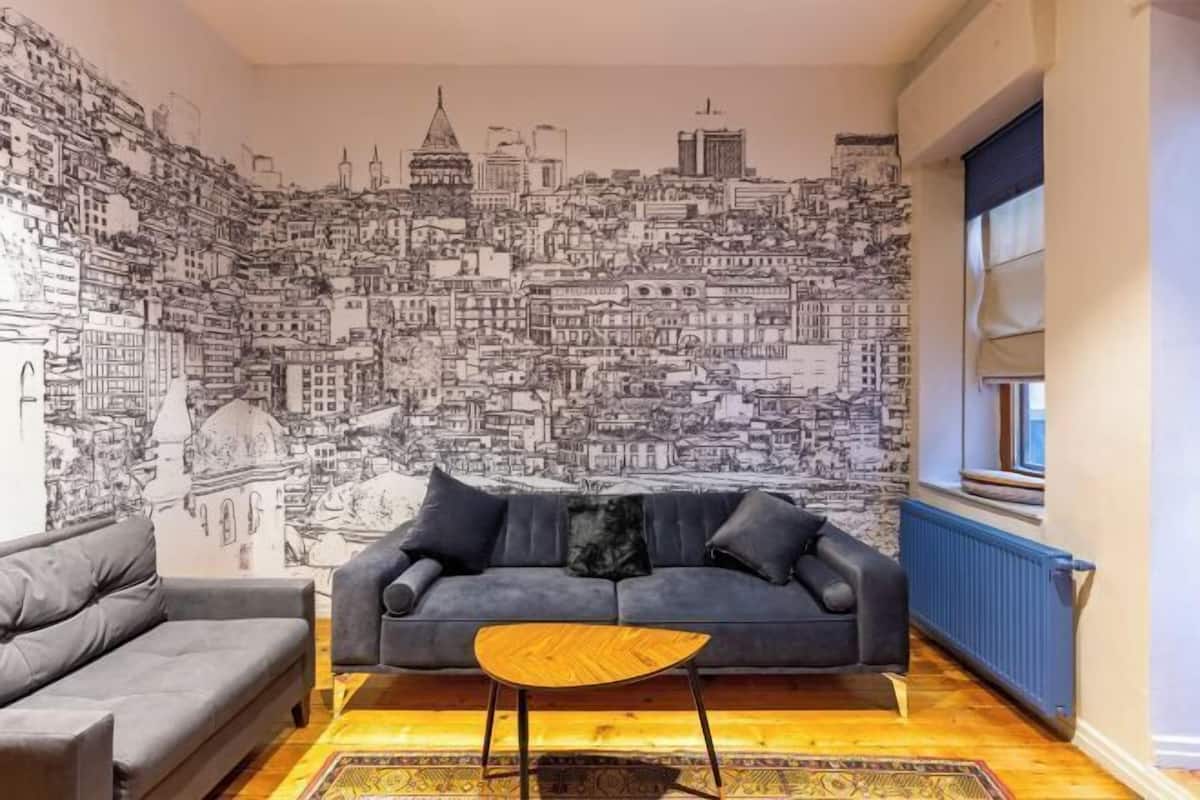 Una sala de estar con un mural de gran tamaño que muestra un paisaje urbano, un sofá gris, un sofá gris más pequeño y una mesa de centro de madera.
