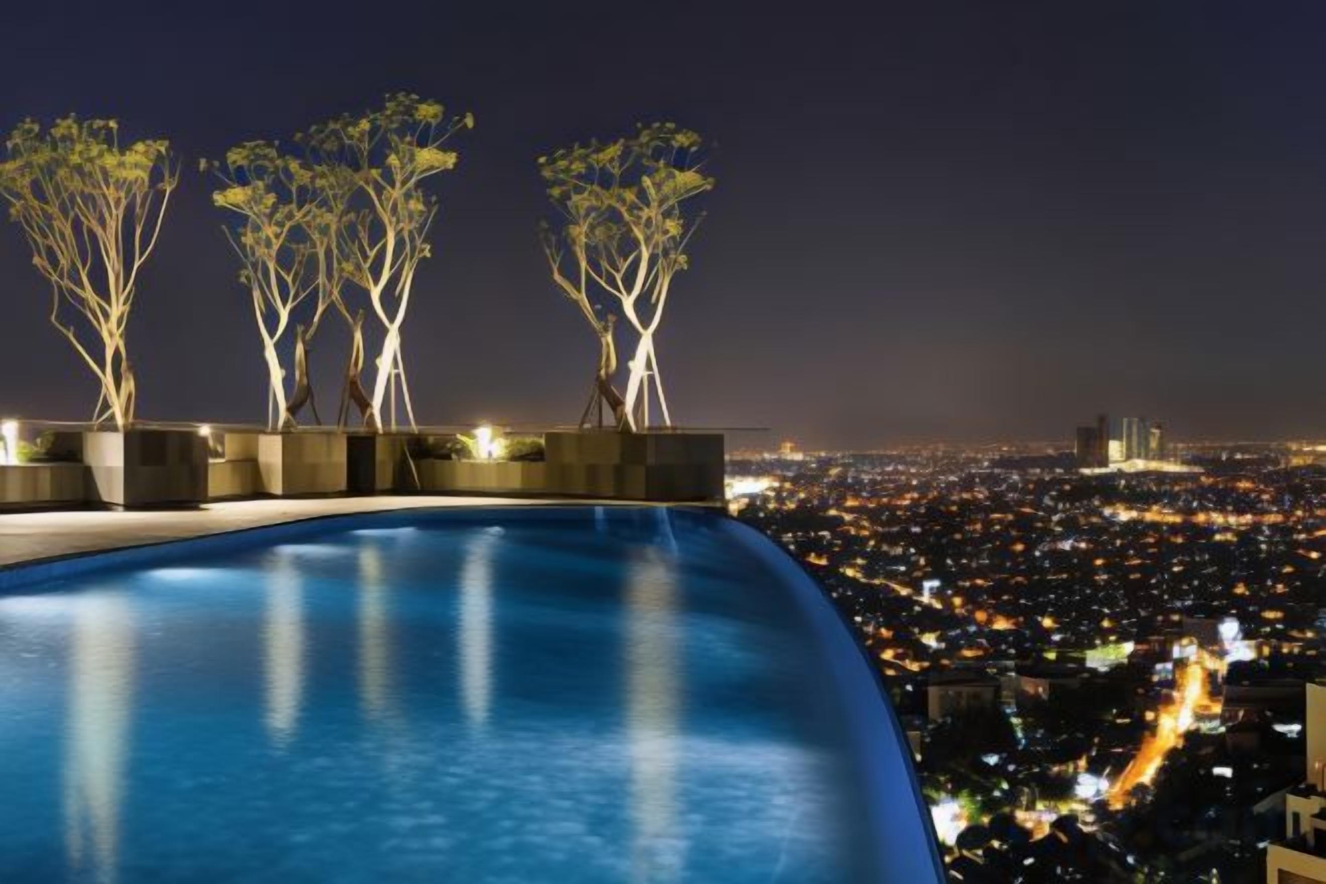 Vista nocturna de una piscina con tres árboles iluminados y un paisaje urbano al fondo.