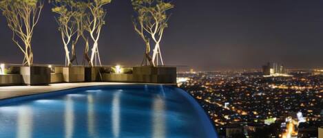 Vista nocturna de una piscina con tres árboles iluminados y un paisaje urbano al fondo.