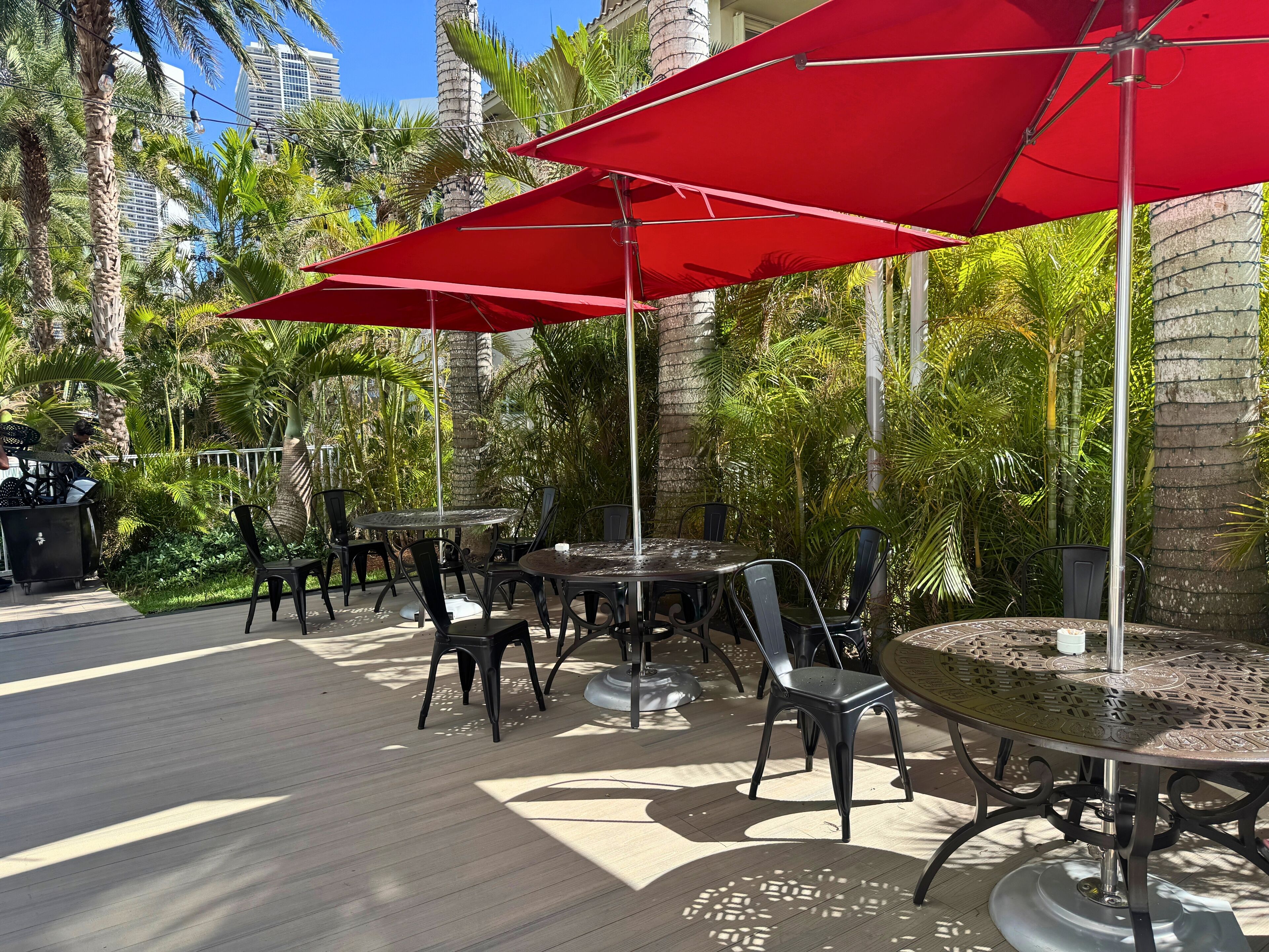An outdoor patio area with tables and chairs, red umbrellas, and palm trees.