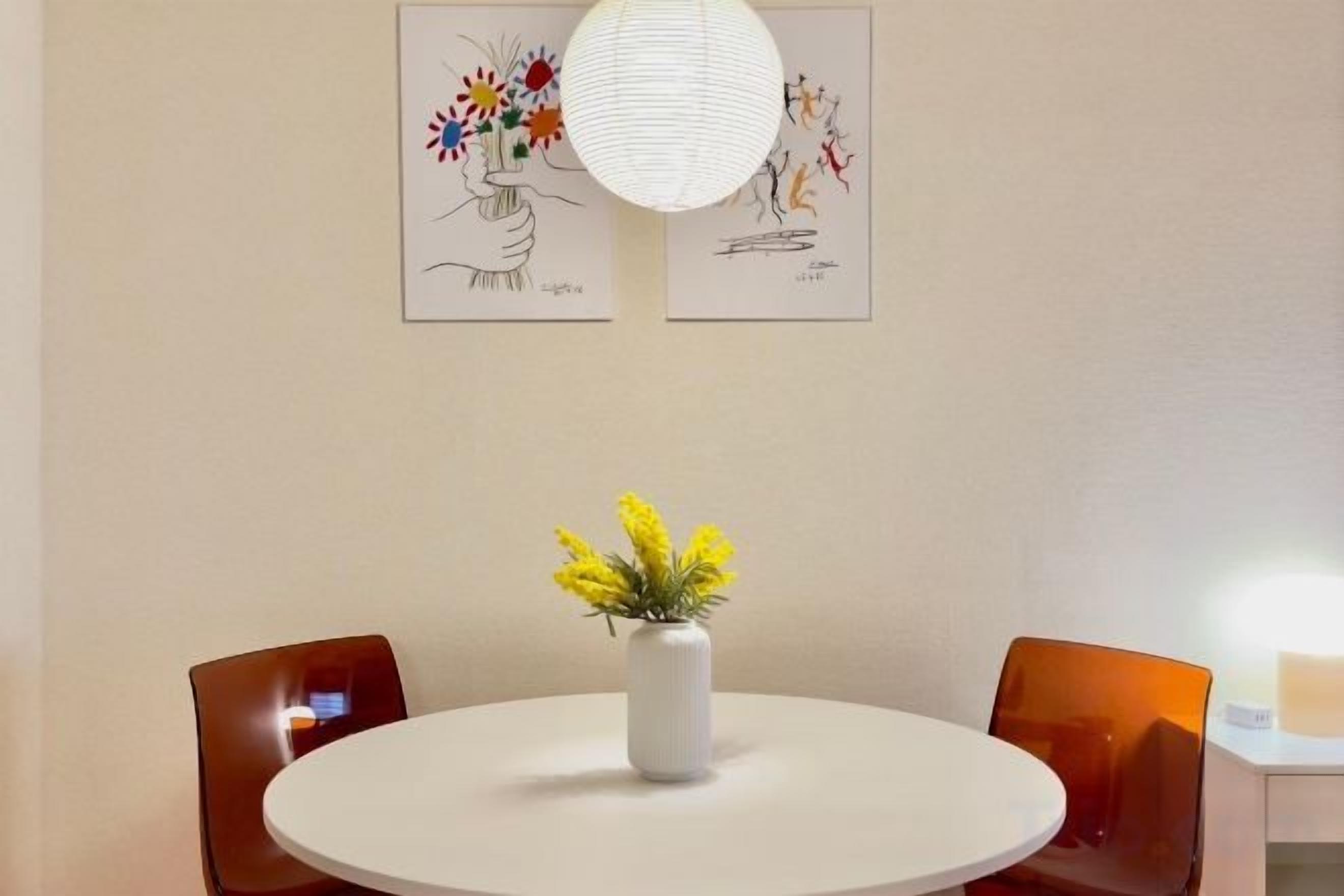 A round dining table with a white vase of yellow flowers, two brown chairs, and two framed artworks on the wall.