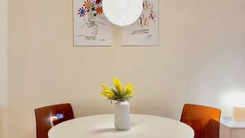 A round dining table with a white vase of yellow flowers, two brown chairs, and two framed artworks on the wall.