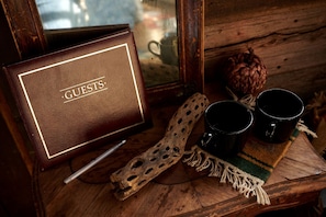 A wooden table with a leather-bound sign reading 'GUESTS,' two black mugs, a carved wooden spoon, and a woven placemat.