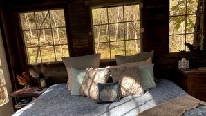 Cama com roupa de cama estampada, almofadas e manta em quarto com paredes de madeira e janelas amplas.