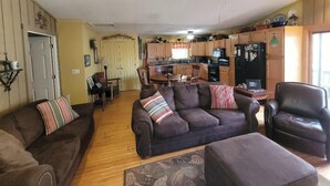 A living room with a brown leather sofa, a brown armchair, and a wooden coffee table. There is a kitchen in the background with wooden cabinets and a refrigerator.
