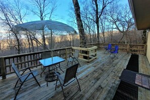 Eine Holzdiele mit Tisch und Stühlen, einem überdachten Bereich und Blick auf Bäume und Hügel.