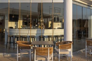 An outdoor seating area with wicker chairs and tables, a bar area with stools, and a large glass window reflecting the interior. - Arkadia Hotel (Arkadii)