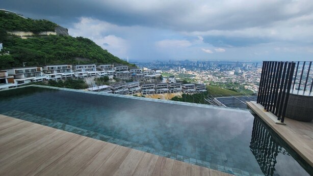 A modern infinity pool with a wooden deck, overlooking a cityscape and a green hillside.