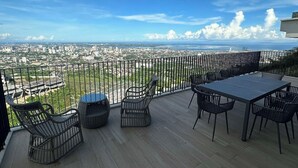 A balcony with wicker furniture and a table, overlooking a cityscape with buildings and a body of water.