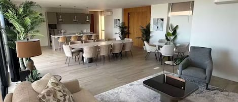 A modern living room with a sofa, coffee table, and chairs. There is a dining area with a table and chairs. The room has a large window and a plant.
