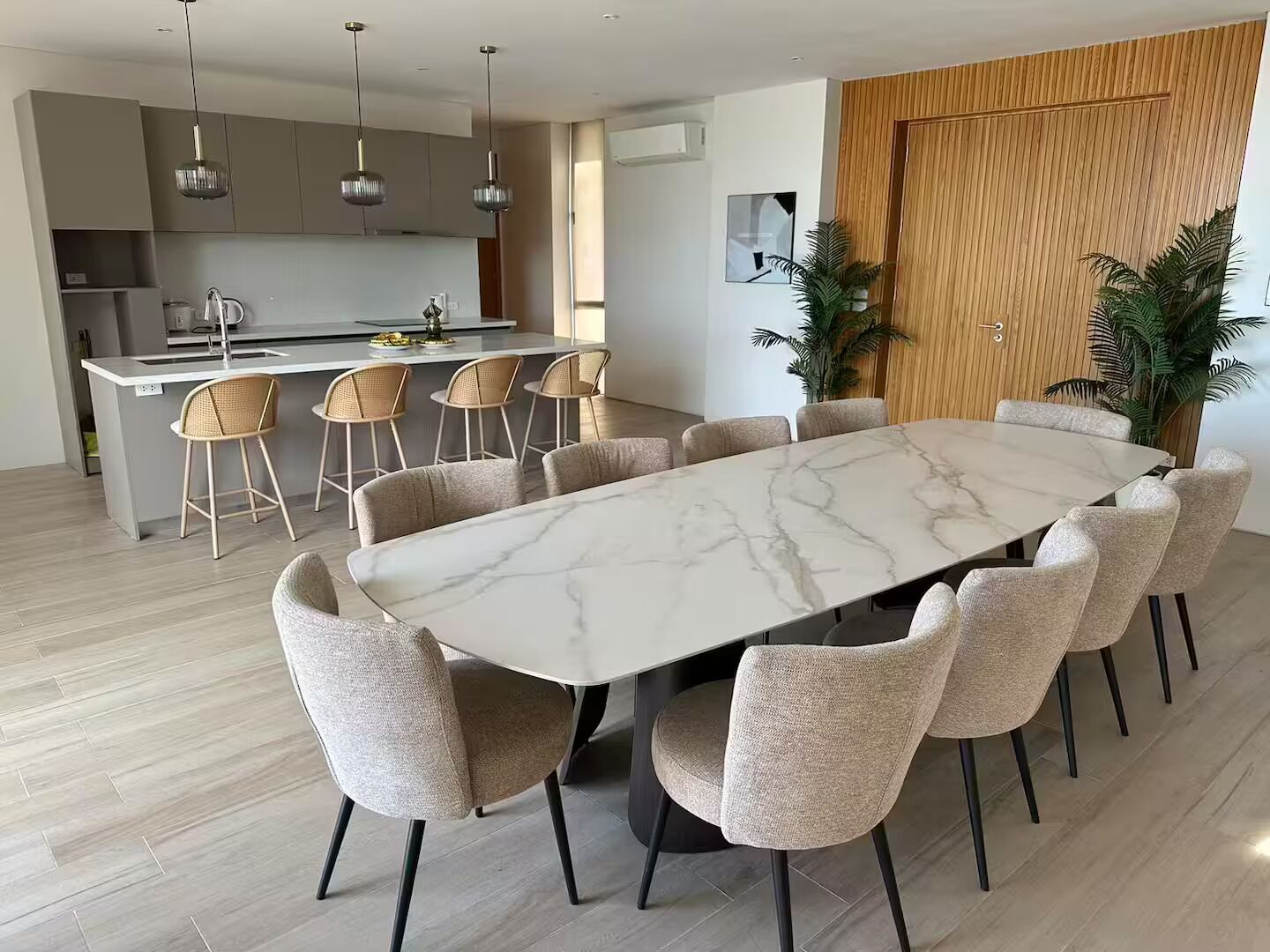 Une cuisine moderne avec des luminaires suspendus, un coin repas doté d’une table en marbre et de chaises rembourrées, et un mur revêtu de panneaux en bois.