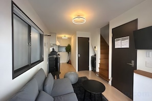 A modern living room with a sofa, a small round table, and a black rug. There is a kitchen area with a stove and cabinets in the background. - URBAN HOUSE NAKANO (Tokyo)
