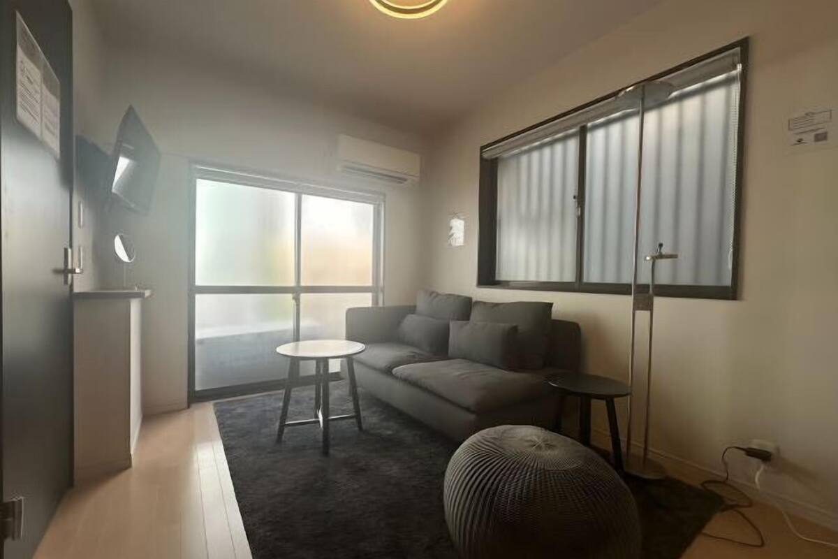 Een moderne woonkamer met een bank, een klein rond tafeltje, een groot raam met jaloezieën en een aan de muur bevestigde televisie.