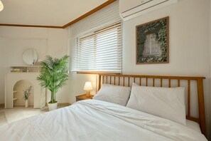 A bedroom with a bed, bedside table, lamp, potted plant, and a painting on the wall.