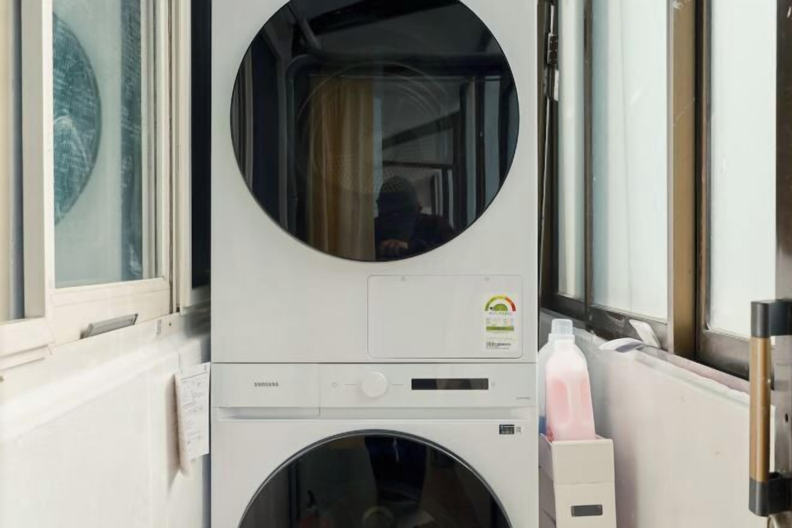 Une machine à laver à chargement frontal, dotée d’une fenêtre circulaire, d’un panneau de commande et d’une bouteille de lessive placée à droite.