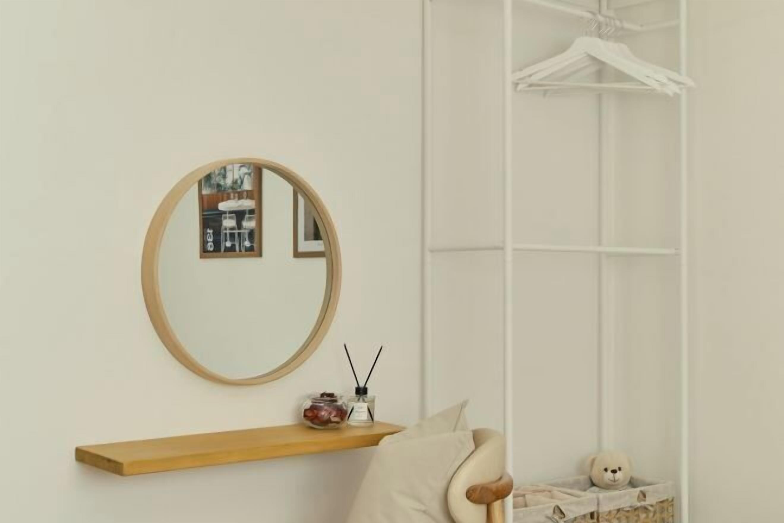 Une pièce au décor minimaliste, avec un miroir rond, une étagère en bois agrémentée d’objets décoratifs, un fauteuil rembourré et une armoire blanche dotée d’une tringle pour suspendre les vêtements.