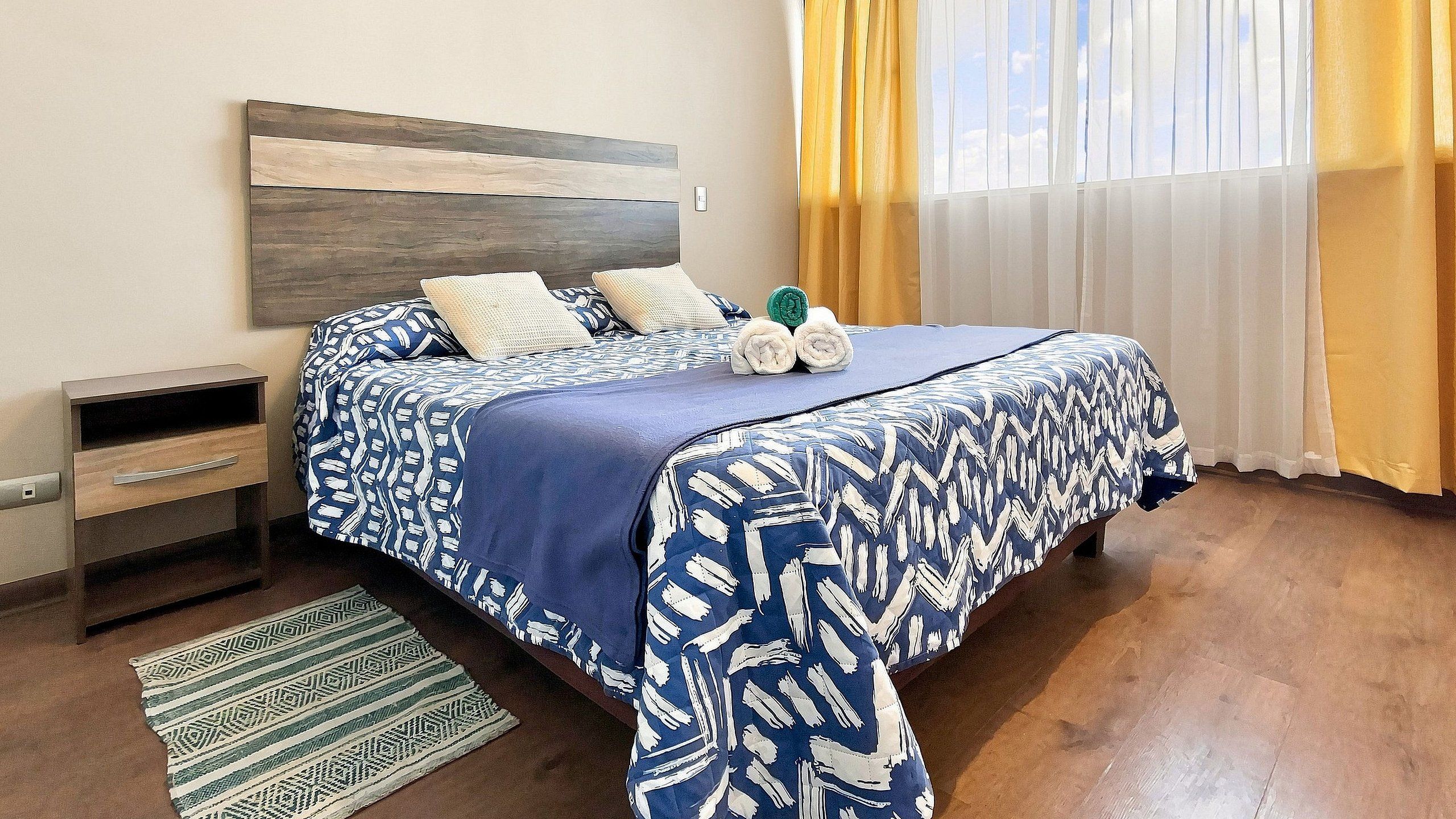 A hotel room with a bed, a nightstand, a lamp, a window with curtains, and a patterned rug.