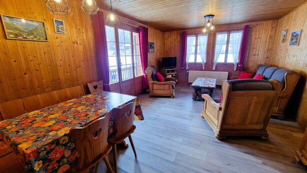 A wooden interior with a dining table, chairs, a sofa, and a television.
