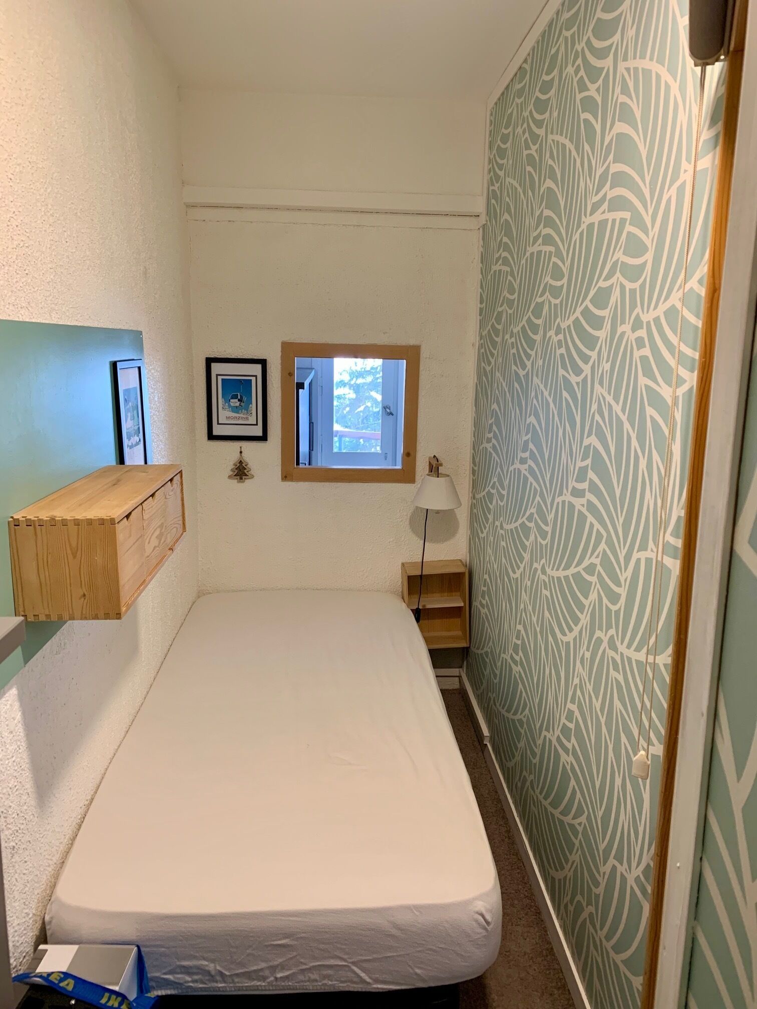 Una habitación pequeña y estrecha con una cama individual, un ventanal, un estante en la pared y una cortina estampada.