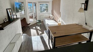 Ein kompakter, gut beleuchteter Raum mit einem Bett, einem kleinen Schreibtisch und einem Stuhl. Zwei Fenster sorgen für natürliches Licht und einen Ausblick ins Freie.
