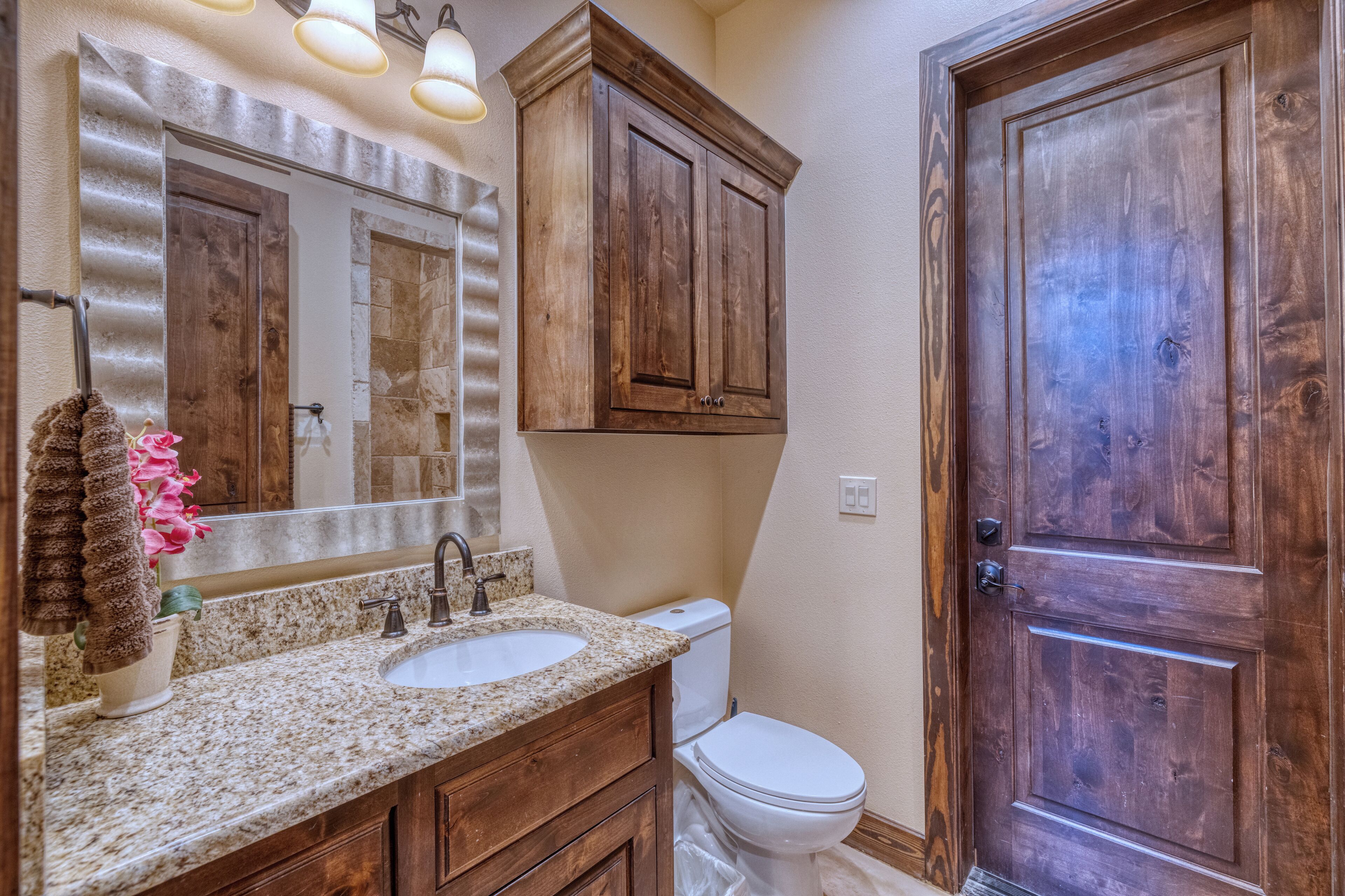 Un baño con lavamanos de encimera de granito, armarios de madera, inodoro blanco y puerta de madera.
