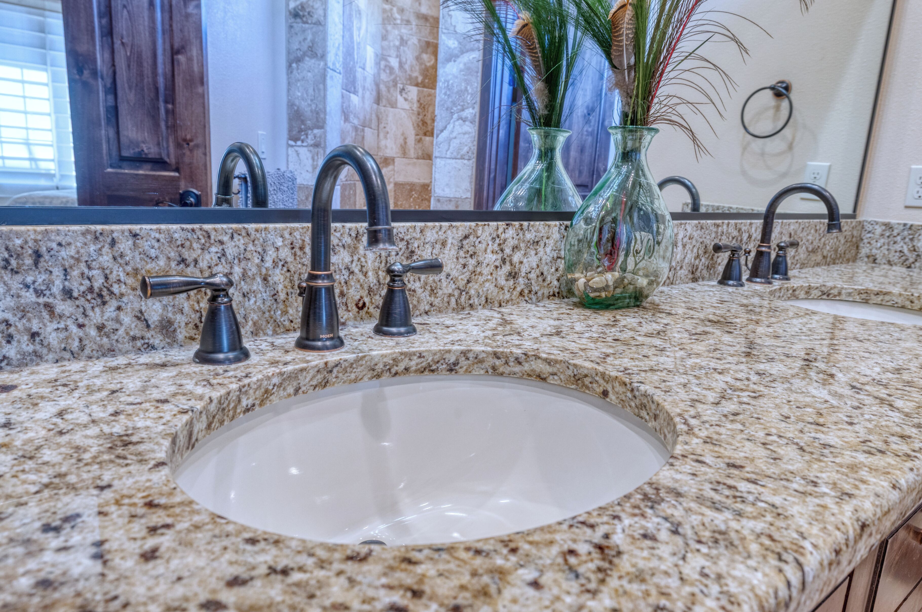 Une salle de bain avec un comptoir en granit, un grand lavabo et deux robinets. Un vase contenant des plantes est posé sur le comptoir.