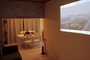 A room with a dining table, chairs, a sofa, and a projector screen displaying a cityscape. - KYOTO TANGO MIYAZU inn (Miyazu)