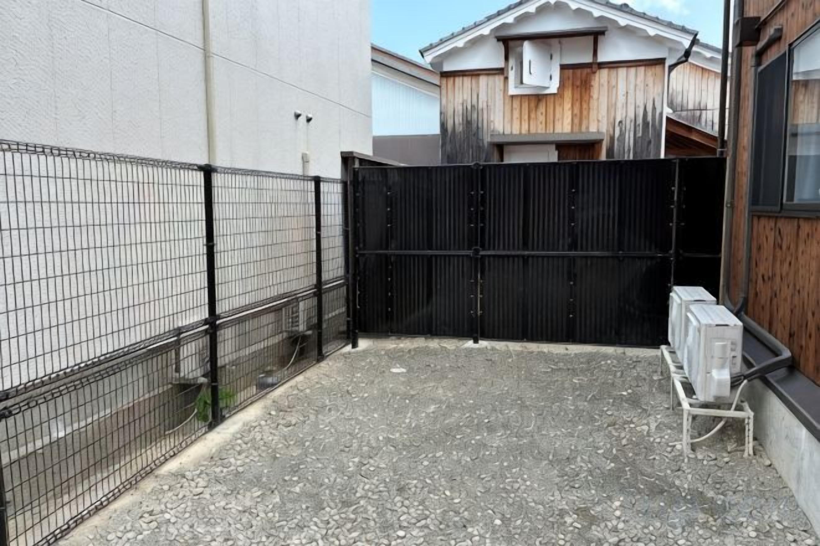 Un área exterior cercada con superficie de grava, una puerta negra y un cuadro de electricidad.