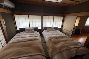A traditional Japanese-style room with tatami flooring, two beds with brown bedding, sliding doors, and a window with a grid pattern.