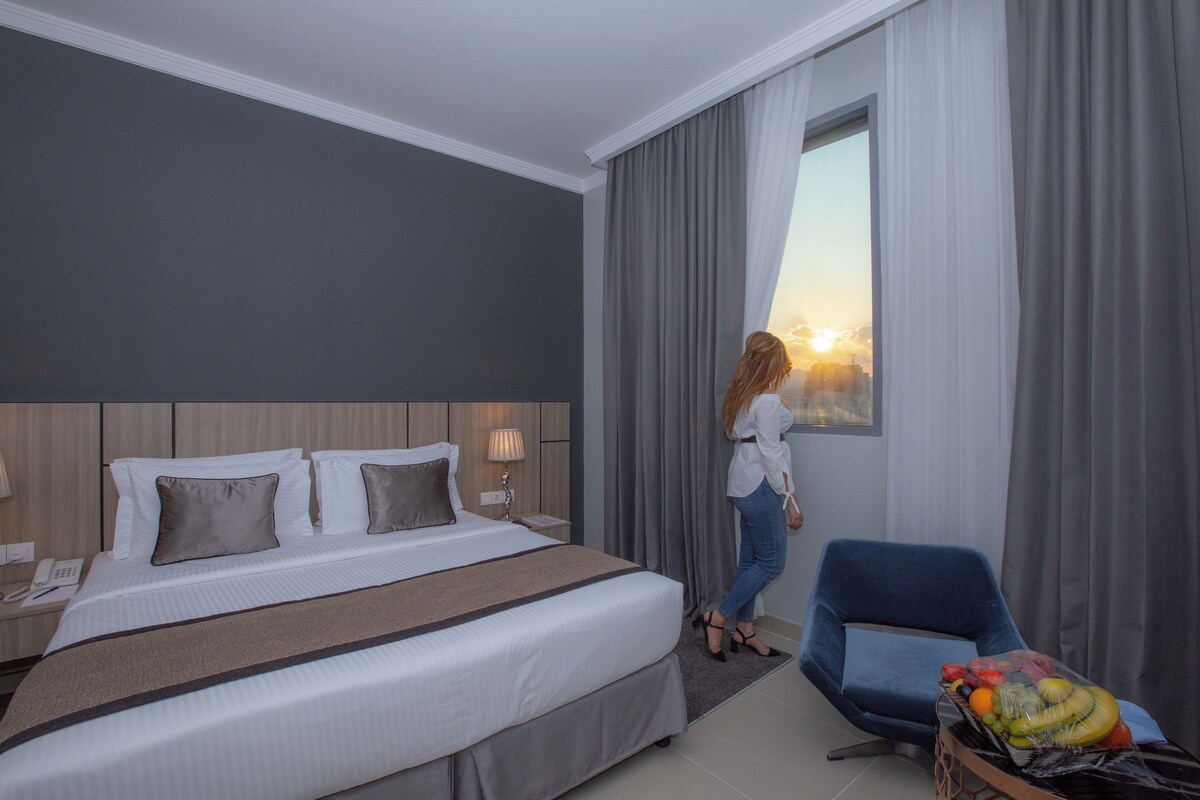 Ein Hotelzimmer mit einem großen Bett, einem Nachttisch, einem Schreibtisch, einem Stuhl und einem Vorhangfenster.