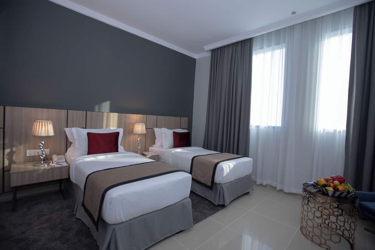 A hotel room with two beds, a nightstand with a lamp, a small table with fruit, and a large window with curtains.
