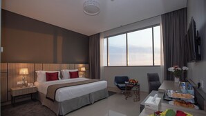 A hotel room with a large bed, bedside tables, a desk with a chair, a TV, and a view of the city. - Fortis Hotel Fujairah (Al-Fujairah)