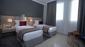 A hotel room with two beds, a nightstand with a lamp, a small table with fruit, and a large window with curtains. - Fortis Hotel Fujairah (Al-Fujairah)