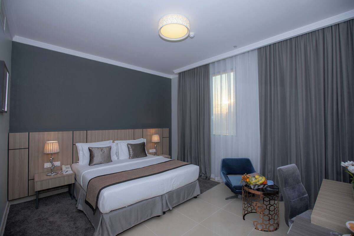 A hotel room with a large bed, a desk with a chair, a small table with a lamp, and a large window with curtains.