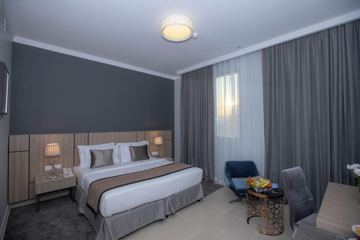 Ein Hotelzimmer mit einem großen Bett, einem Schreibtisch mit Stuhl, einem kleinen Tisch mit Lampe und einem großen Fenster mit Vorhängen.