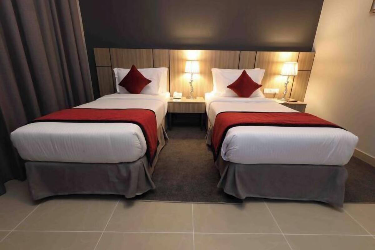 A hotel room with two beds, each with a red blanket, a wooden headboard, bedside lamps, and a dark curtain.