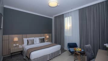 A hotel room with a large bed, a desk with a chair, a small table with a lamp, and a large window with curtains.