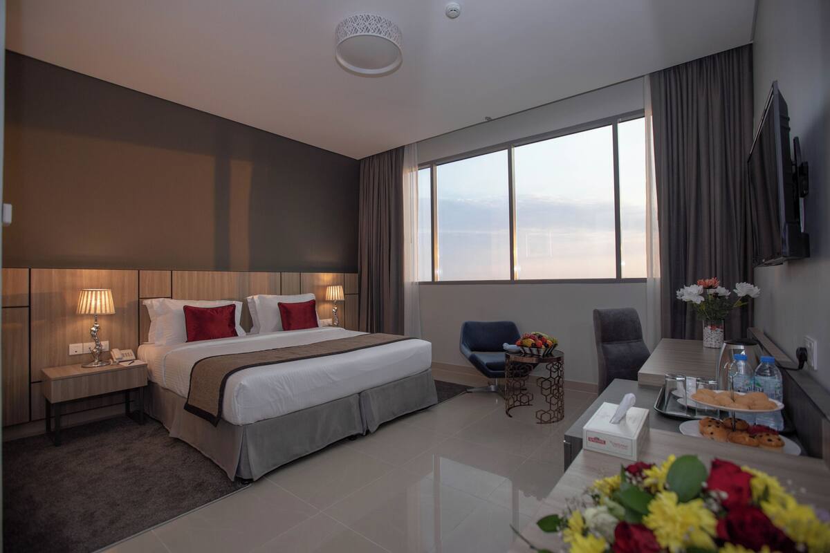 A hotel room with a large bed, bedside tables, a desk with a chair, a TV, and a view of the city.