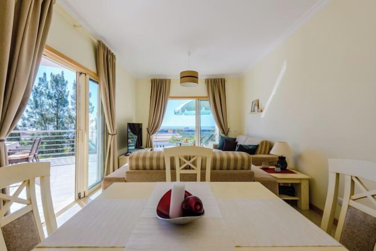 Habitación de hotel con balcón, sofá, mesa de comedor y vistas al exterior.