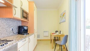 A compact kitchen with a stove, microwave, and coffee maker, a dining area with a table and chair, and a window with a view. - BnB Homes Ocean View Apartment with Huge Sunny Terrace, 2 Swimming Pools & Tennis Court (Albufeira)