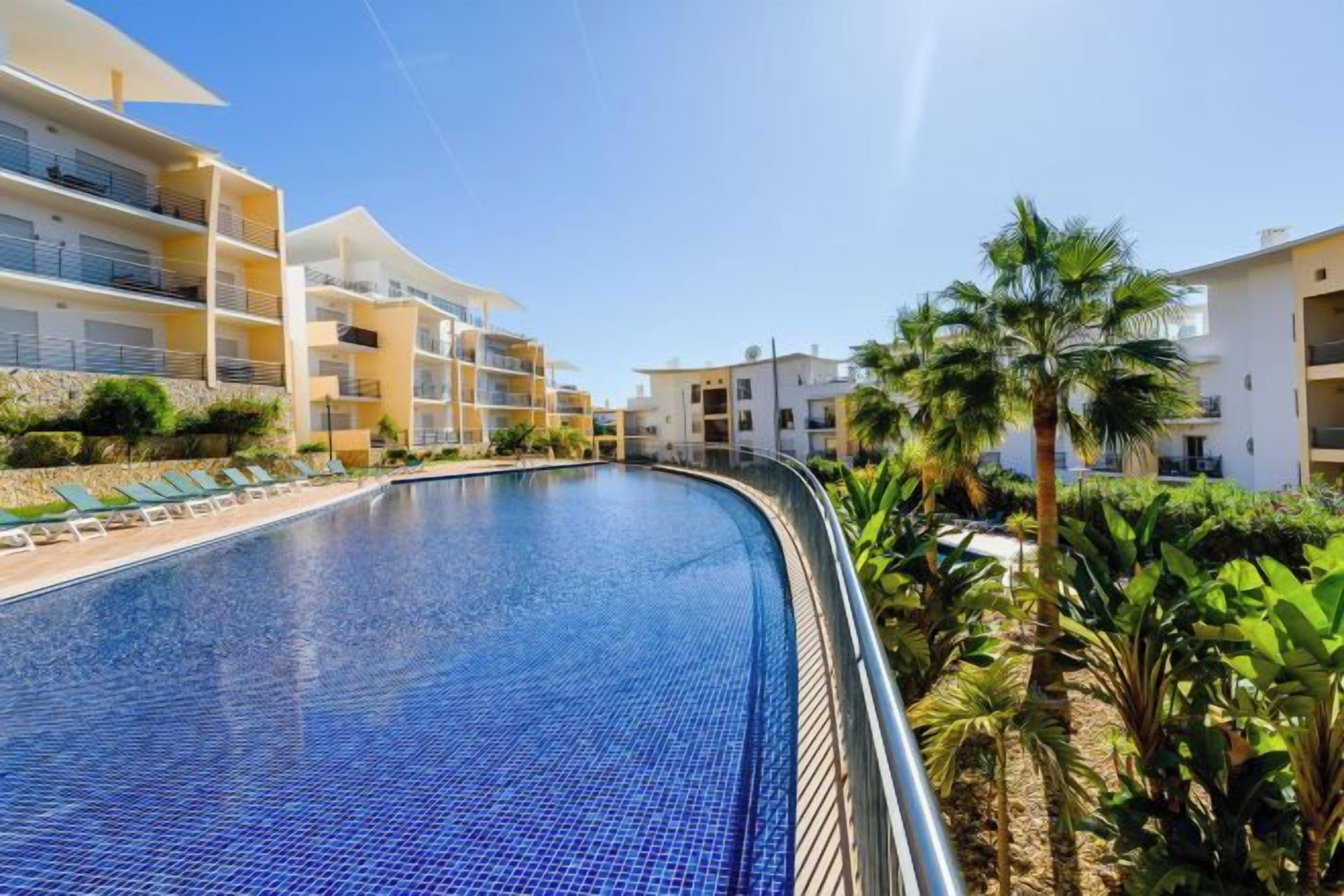 A swimming pool with a curved edge, surrounded by lush greenery and multi-story buildings with balconies.