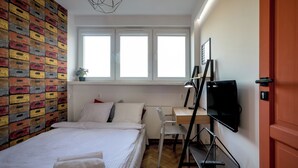 A bedroom with a bed, a desk with a computer, a TV, and a window with blinds. - RentPlanet - Apartament plac Nowy Targ (Wrocław)
