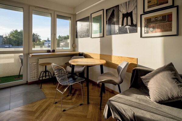 Ein gemütliches Zimmer mit Holzboden, einer Couch, einem Esstisch und Stühlen. Große Fenster bieten einen Blick ins Freie.