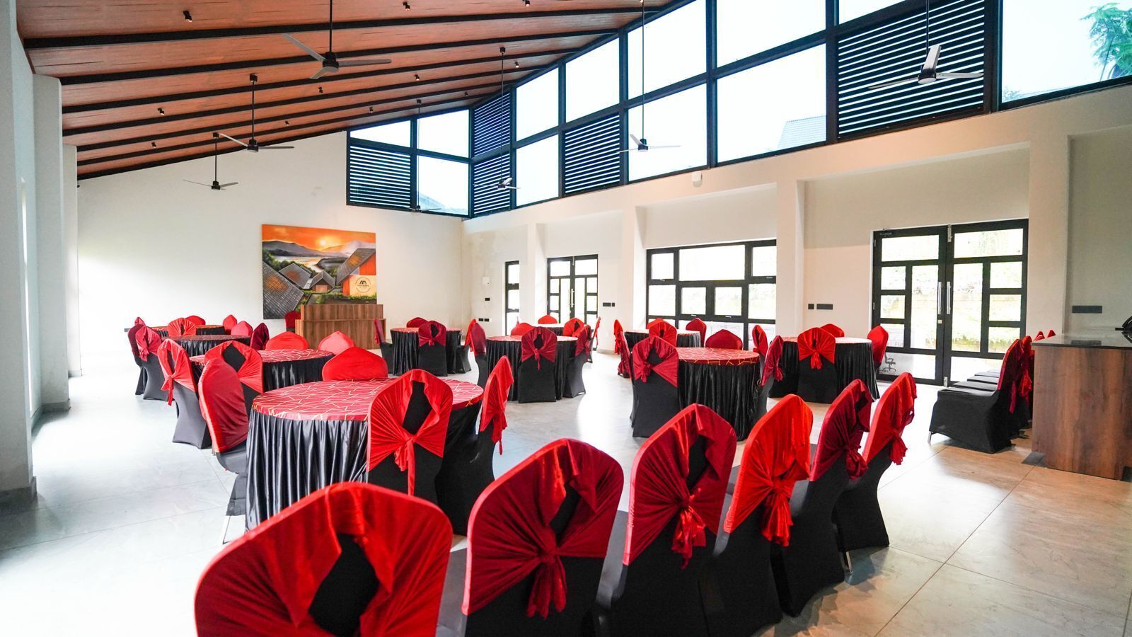 A spacious hall with multiple round tables, each covered with black tablecloths and red chair covers. The room has large windows allowing natural light and a high ceiling with exposed beams.