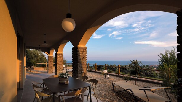 Pátio externo com mesa e cadeiras, pilares de pedra e vista para o mar.