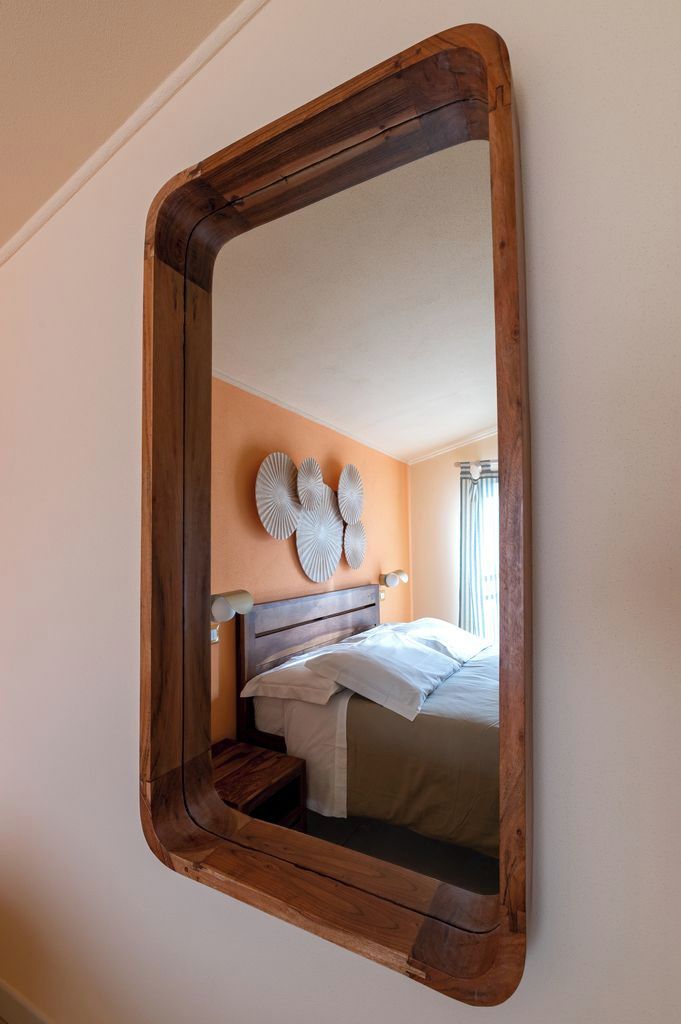 Camera da letto con uno specchio con cornice in legno, un letto con lenzuola bianche, un comodino e un punto luce a parete.