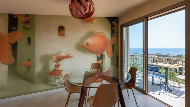 Un comedor con mesa y sillas de vidrio, un mural con motivos de peces en la pared y un balcón con vistas al mar.