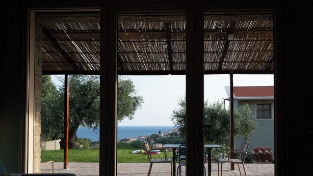 Una habitación con un gran ventanal corredizo que da a un patio exterior con mesa y sillas, vista al mar y un edificio al fondo.