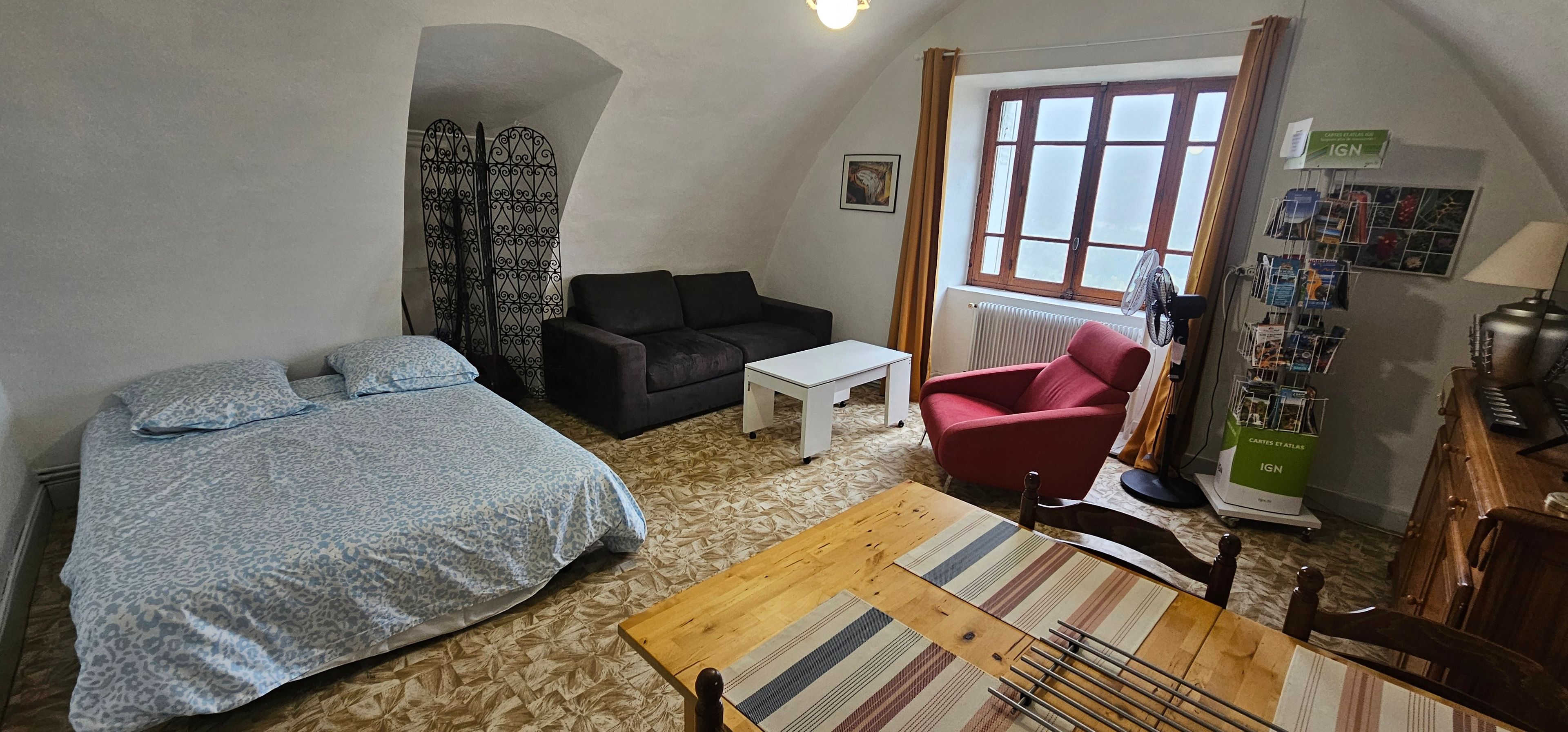Une petite pièce agréable et chaleureuse, avec un lit, un canapé, un fauteuil rouge, une table en bois et un radiateur.